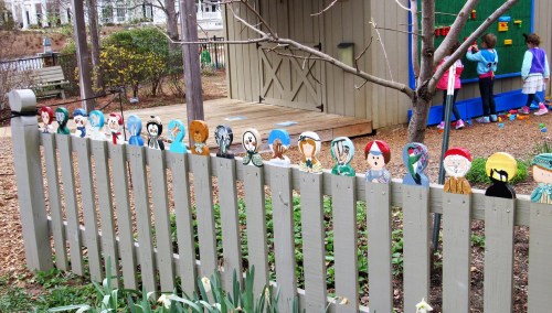 Colorful wooden figures line a wooden fence in a park, with children playing in the background near a play structure.