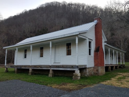 A white, one-story house with a metal roof, set on stone pillars, surrounded by grass and mountains in the background.