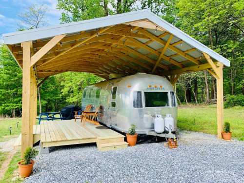 A silver Airstream trailer parked under a wooden shelter, surrounded by greenery and gravel, with potted plants nearby.