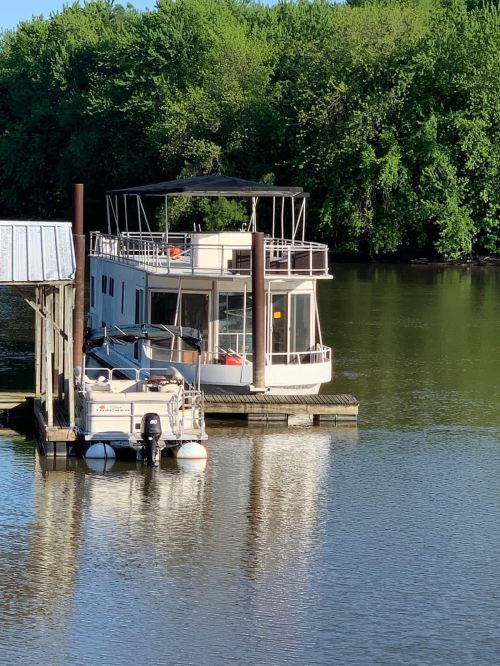 A houseboat docked at a pier on a calm river, surrounded by lush green trees.