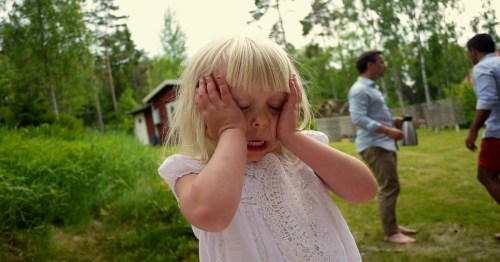 A young child with blonde hair covers their face with hands, looking distressed, while two adults are blurred in the background.