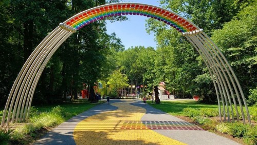 A colorful rainbow arch over a path leading to a playground, surrounded by lush green trees.