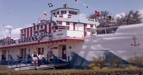 A colorful building resembling a ship, decorated with flags, with children playing outside in a garden setting.