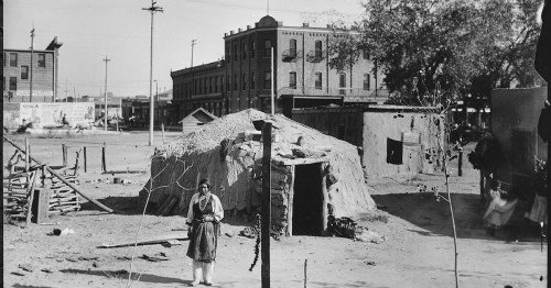 A historical black-and-white photo of a woman standing outside a traditional adobe structure in a town setting.