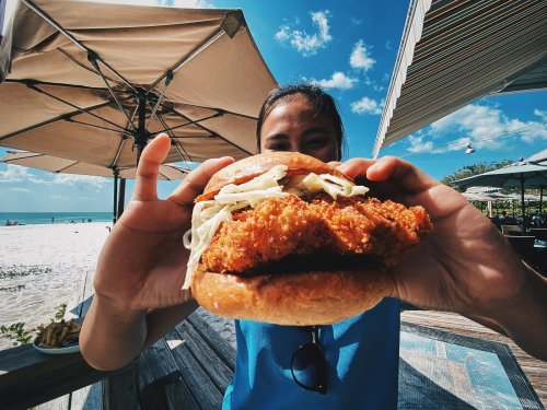 A person holds a large fried chicken sandwich with lettuce, against a beach backdrop and clear blue sky.