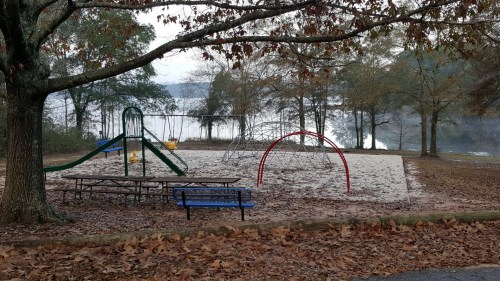 A playground with a slide, climbing structure, and swings, surrounded by trees and a lake in the background.