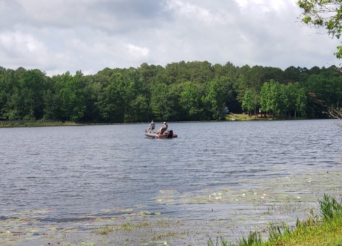 Two people in a small boat fishing on a calm lake surrounded by lush green trees and cloudy skies.