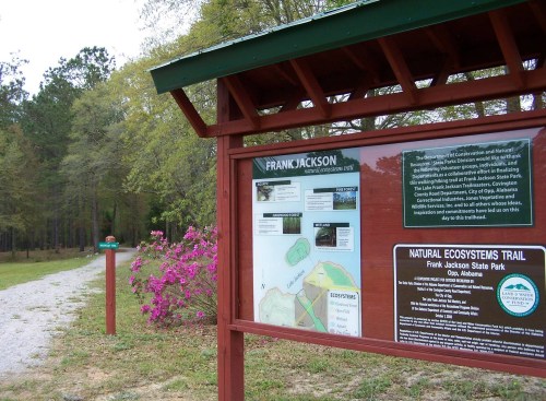 Sign at Frank Jackson State Park displaying a map and information about the Natural Ecosystems Trail, surrounded by greenery.