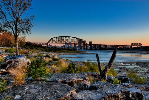A scenic view of a bridge over a river at sunset, with rocky shores and trees in the foreground.