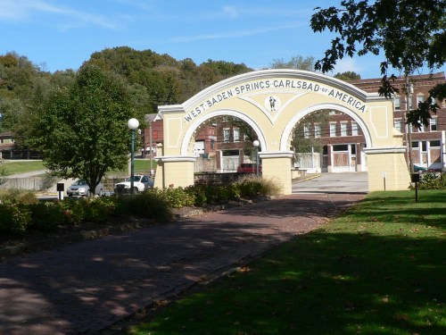 Archway entrance to West Baden Springs, surrounded by trees and a pathway, with historic buildings in the background.