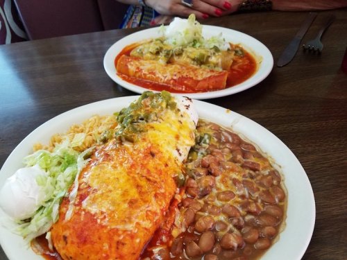Two plates of Mexican food featuring enchiladas, rice, beans, and toppings like cheese and lettuce on a wooden table.