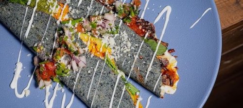 A blue corn tortilla filled with colorful toppings, drizzled with sauce, served on a blue plate.