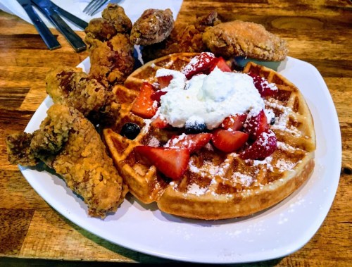 A plate of chicken and waffles topped with strawberries and whipped cream, served with crispy fried chicken pieces.