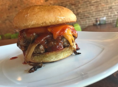 A close-up of a juicy burger with melted cheese, grilled onions, and sauce, served on a toasted bun on a white plate.