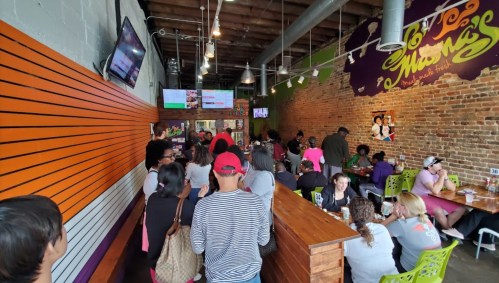 A busy restaurant interior with patrons dining and ordering, featuring colorful decor and a brick wall.