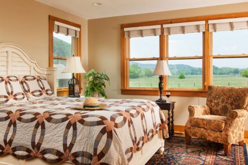Cozy bedroom with a quilted bed, armchair, and large windows showcasing a scenic view of green hills.