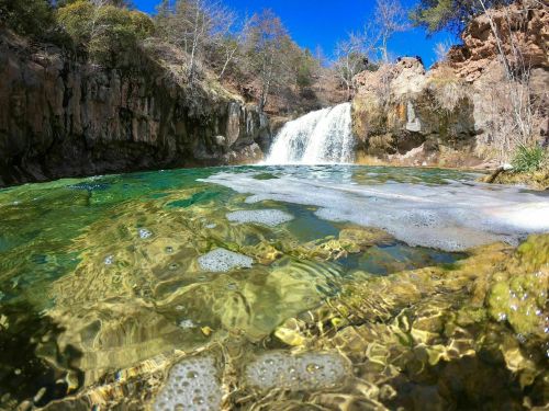 A serene waterfall cascades into a clear, green pool surrounded by rocky cliffs and trees under a bright blue sky.