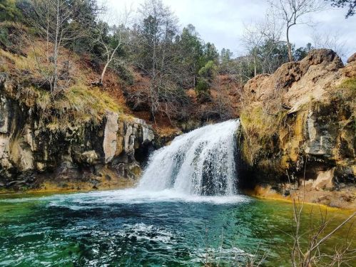 A serene waterfall cascades into a clear pool, surrounded by rocky cliffs and lush greenery.