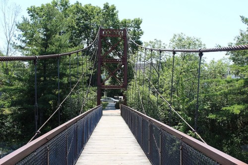 A suspension bridge with a wooden walkway surrounded by lush green trees on a sunny day.