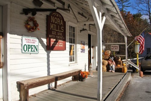 A rustic storefront with signs reading "Open" and "Nora Mills," featuring autumn decorations and teddy bears on the porch.
