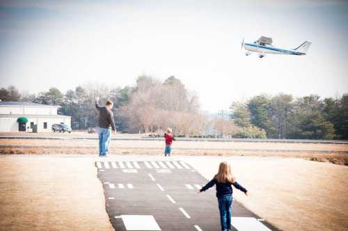 A man and two children wave as a small plane flies overhead near a runway.