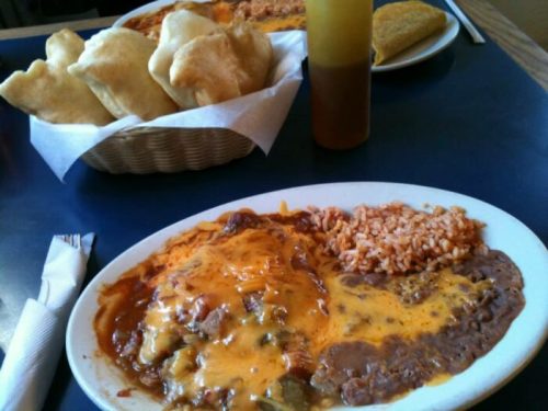 A plate of cheesy beef enchiladas with rice and refried beans, accompanied by a basket of fried pastries.