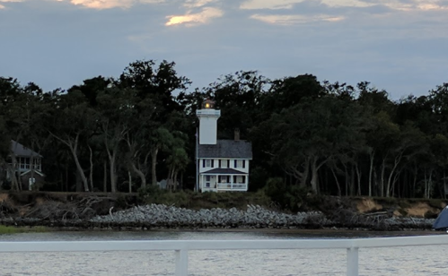 A lighthouse stands on a shore surrounded by trees, with a cloudy sky above and water in the foreground.