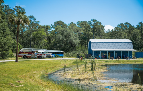 A blue building by a pond, with a fire truck and a bus parked nearby, surrounded by trees and grass.