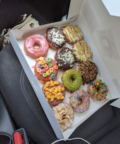 A box of colorful donuts with various toppings, including sprinkles, cereal, and chocolate, resting on a car seat.