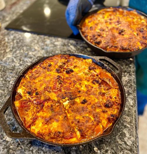 Two cast iron pans of baked lasagna on a granite countertop, one being held with an oven mitt.
