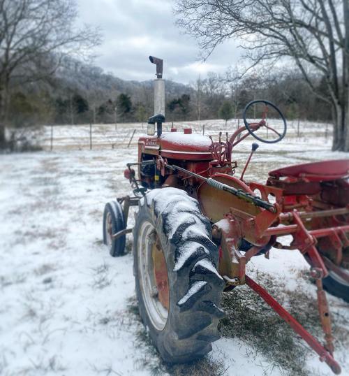 A red tractor covered in snow, parked in a snowy field with trees and hills in the background.