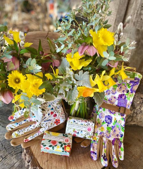 Colorful gardening gloves beside a jar of vibrant flowers and soap bars on a wooden surface.