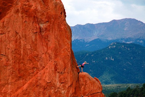 A climber scales a tall, red rock formation with mountains in the background under a cloudy sky.