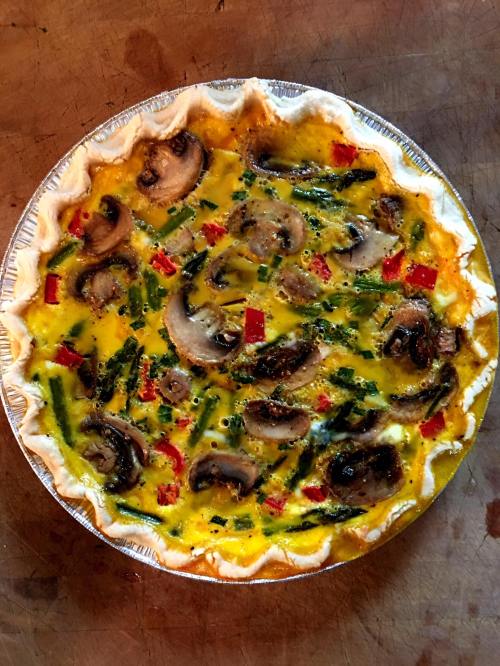 A baked quiche with mushrooms, red peppers, and green onions in a golden crust, placed on a wooden surface.