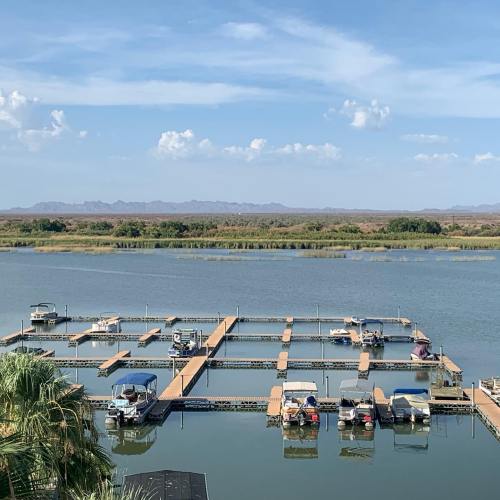 A serene view of a marina with several boats docked, surrounded by calm water and distant mountains under a blue sky.