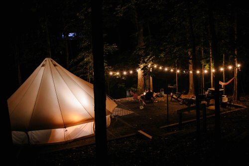 A cozy tent illuminated at night, surrounded by trees and string lights, with a campsite setup nearby.
