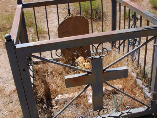 A weathered grave surrounded by a metal fence, overgrown with dry grass and weeds.