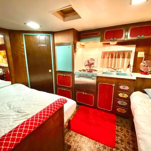 Cozy retro-style room with two beds, a kitchenette, and colorful wooden cabinetry. Red accents and a plush rug add warmth.