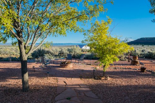 A serene outdoor space with a stone path, fire pit, and trees, overlooking a scenic landscape under a clear blue sky.