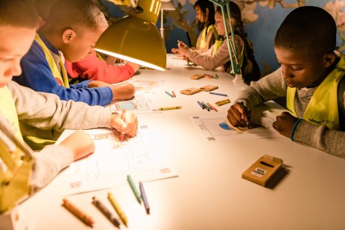 Children in bright vests coloring on paper at a table, illuminated by a lamp, with colored pencils scattered around.