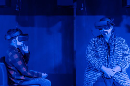 Two people wearing VR headsets sit in a dimly lit blue room, engaged in a virtual experience.