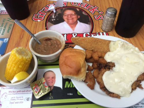 A plate of fried fish, shrimp, a roll, corn, and beans, with a side of creamy sauce, on a restaurant table.