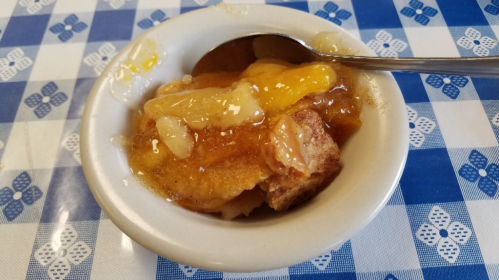 A small bowl of peach cobbler with a spoon, sitting on a blue and white checkered tablecloth.