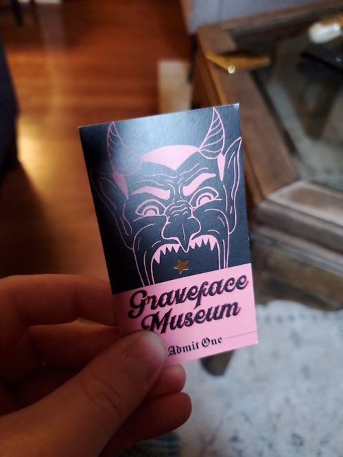 A hand holding a ticket for the Graveface Museum featuring a devilish face design.
