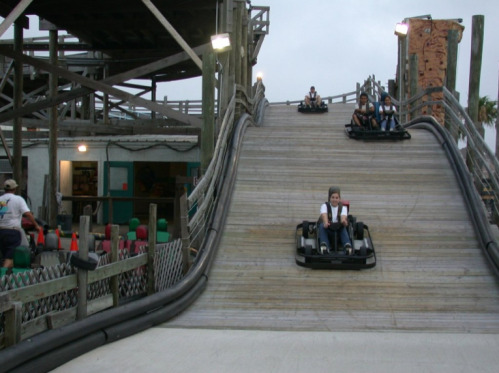 Two go-karts racing down a wooden track at an amusement park, with spectators and a building in the background.