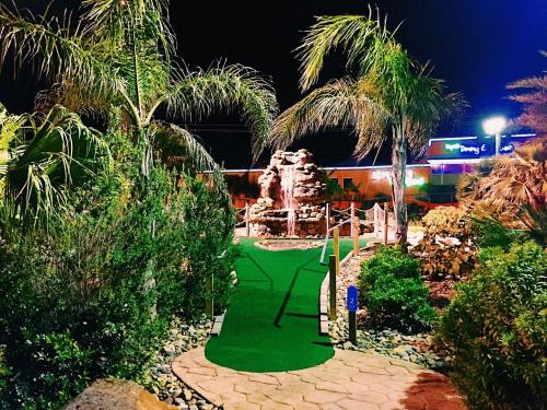 A mini-golf course at night, featuring a green path, palm trees, and a decorative rock fountain.