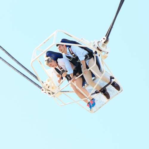 Two people in a harnessed cage, suspended high above, smiling and enjoying a thrilling ride against a clear blue sky.