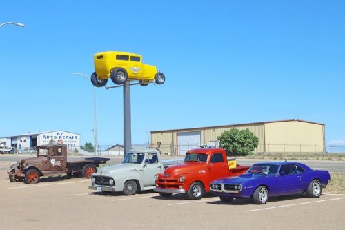A collection of vintage cars, including a yellow hot rod on a pole, parked in a lot with an auto repair shop in the background.