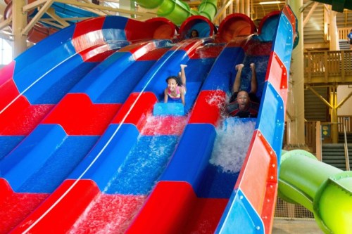 Two children joyfully sliding down colorful water slides at an indoor water park. Water splashes around them.