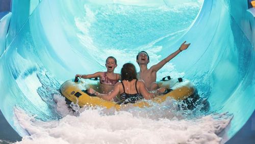 Three children joyfully ride a yellow inflatable tube down a water slide, splashing through waves.